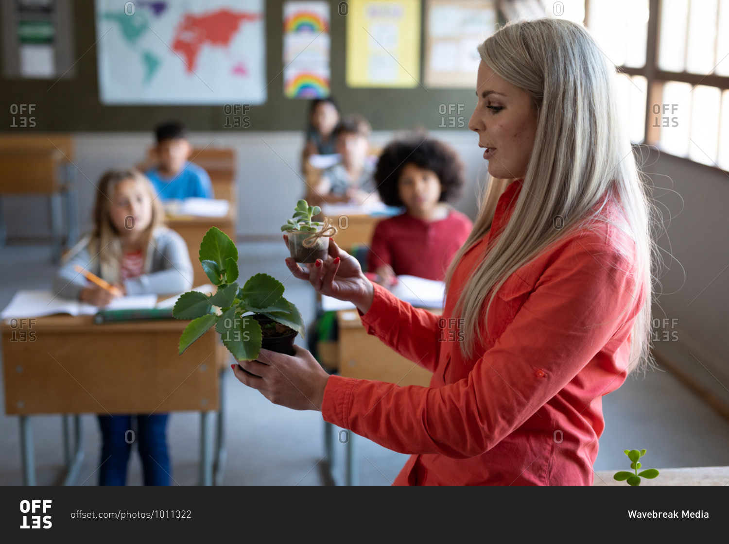 Female Caucasian teacher showing a plant pot to group of multi ethnic kids in class at school. Primary education social distancing health safety during Covid19 Coronavirus pandemic.