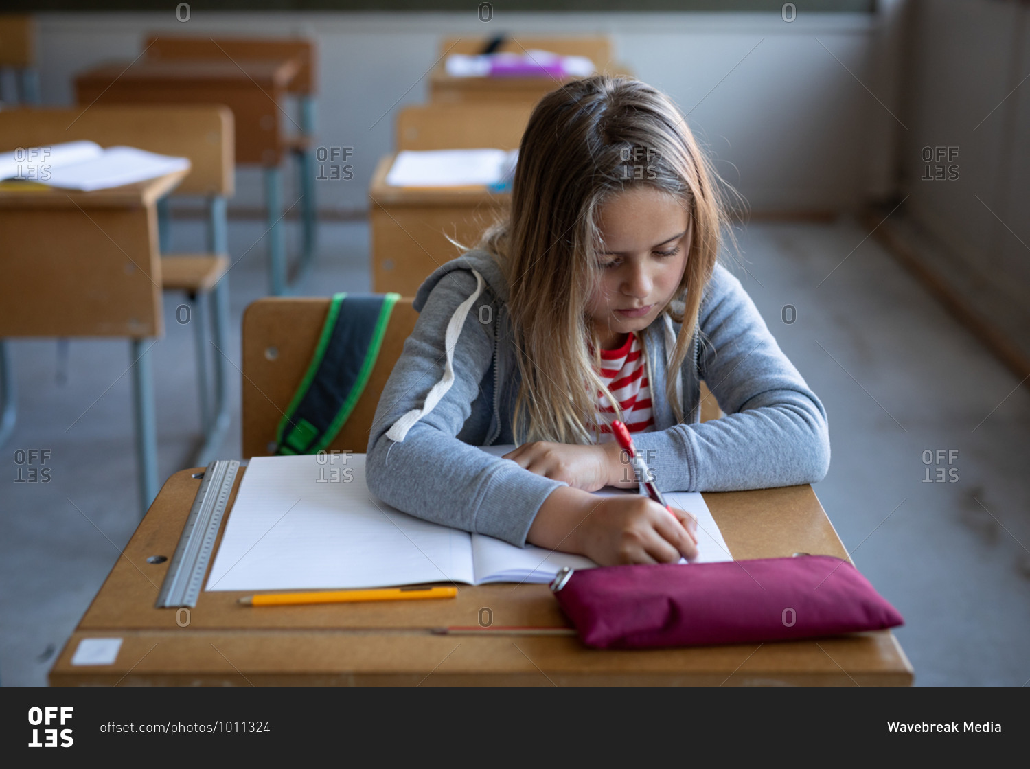 Caucasian girl writing in a book while sitting on her desk at school. Primary education social distancing health safety during Covid19 Coronavirus pandemic.
