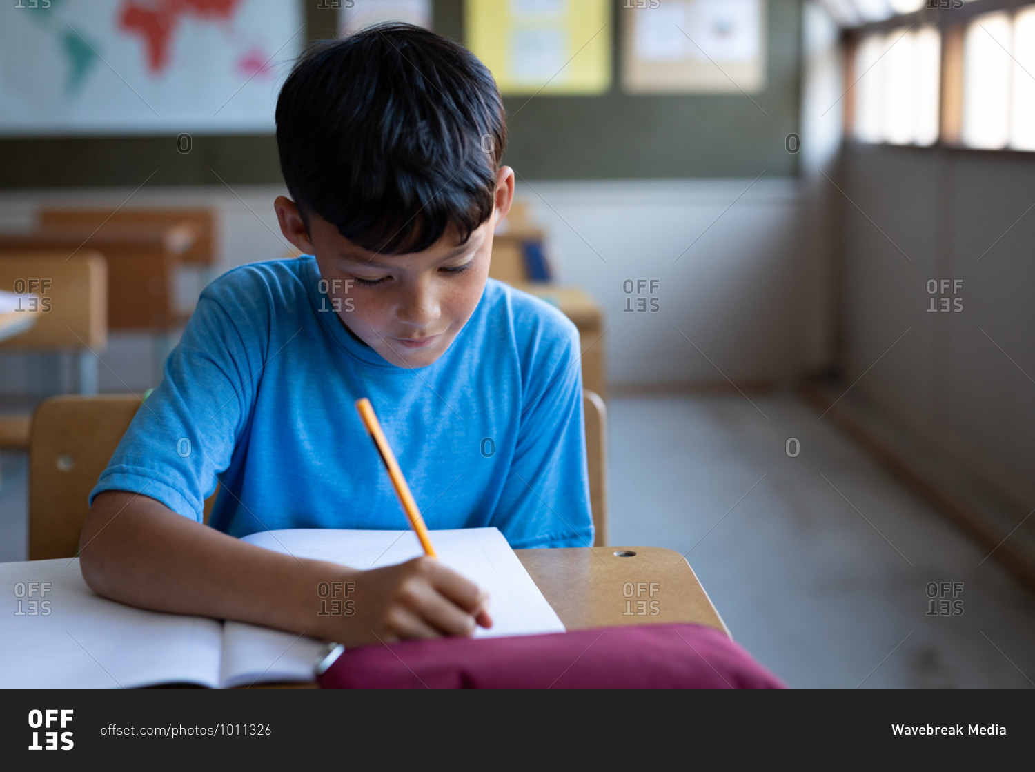 Mixed race boy writing in a book while sitting on his desk at school. Primary education social distancing health safety during Covid19 Coronavirus pandemic.