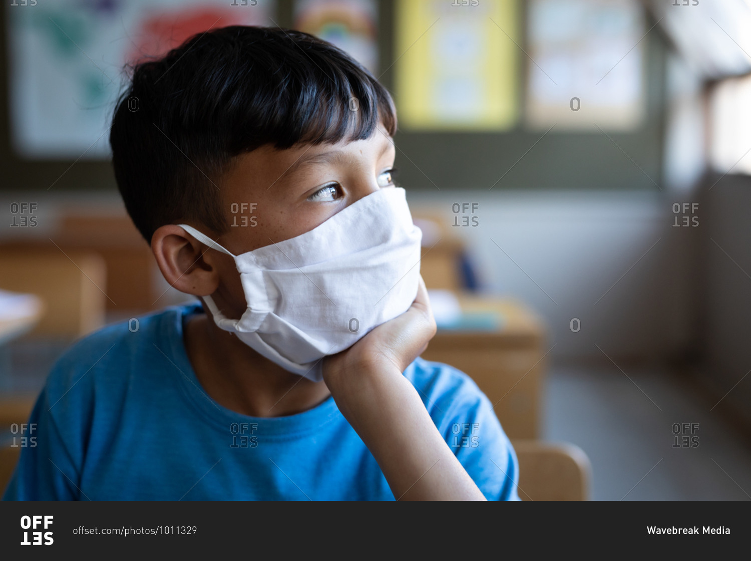 Thoughtful mixed race boy wearing face mask sitting on his desk at school. Primary education social distancing health safety during Covid19 Coronavirus pandemic.
