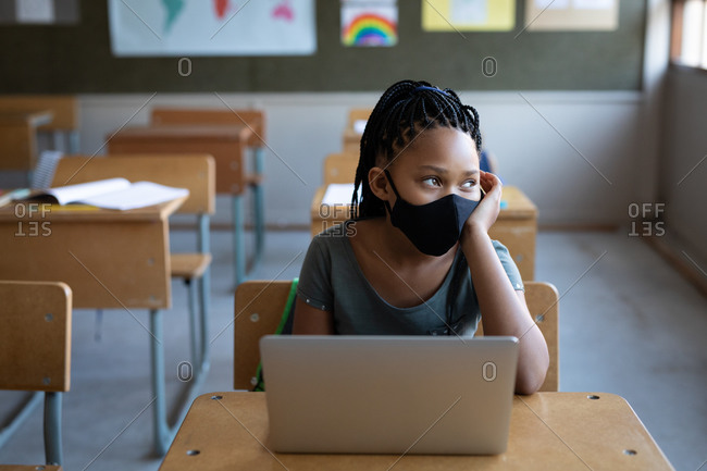 Mixed race girl wearing a face mask, using laptop while sitting on his desk in class at school. Primary education social distancing health safety during Covid19 Coronavirus pandemic.