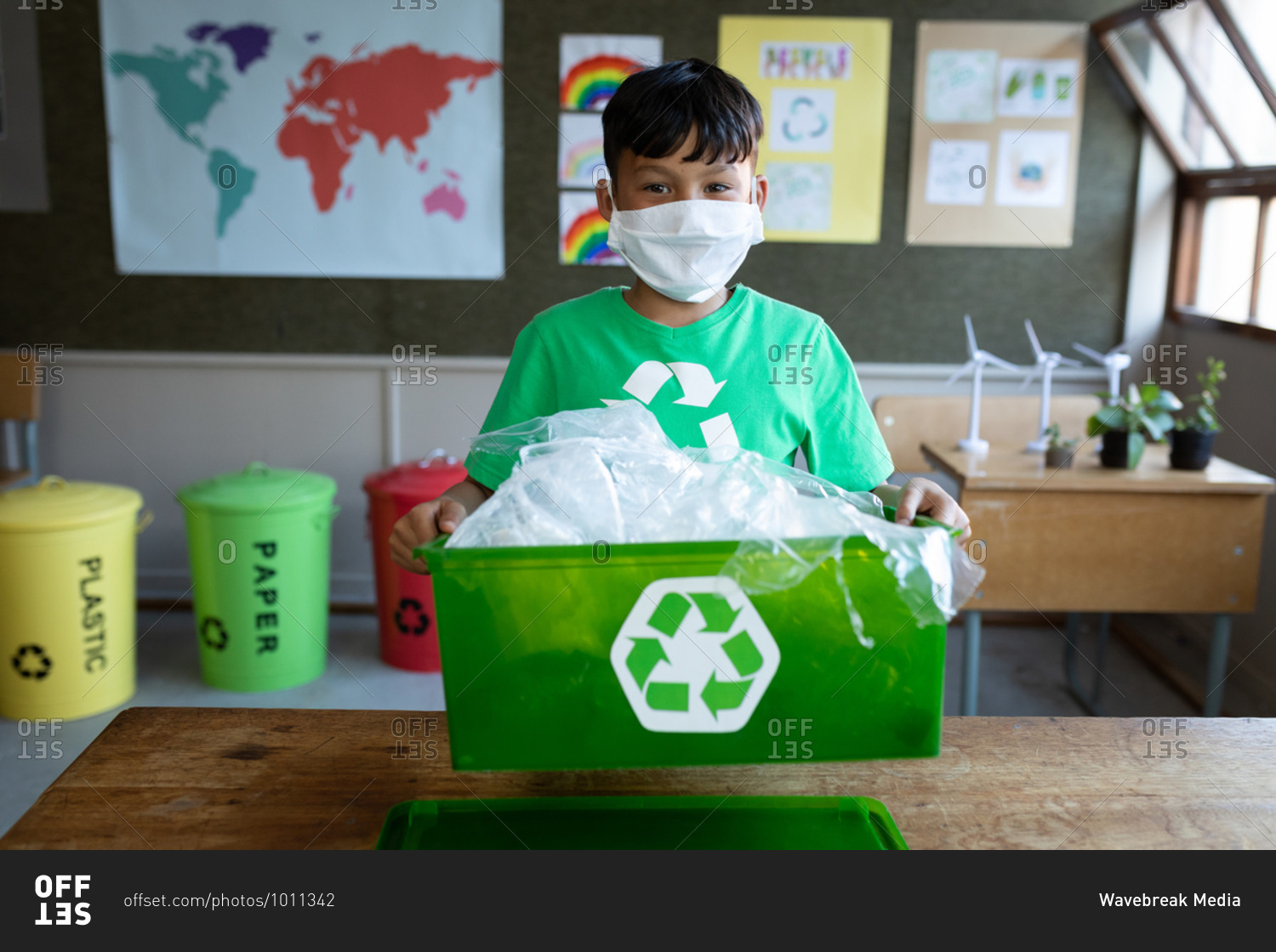 Portrait of a mixed race boy wearing face mask holding a recycle container in class at school. Primary education social distancing health safety during Covid19 Coronavirus pandemic.