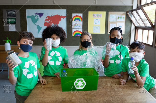 Portrait of a group of multi ethnic kids wearing face masks holding plastic items in class at school. Primary education social distancing health safety during Covid19 Coronavirus pandemic.