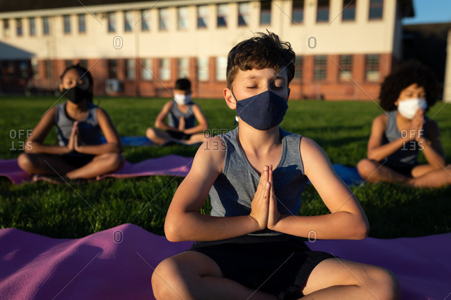 Group of multi ethnic kids wearing face masks performing yoga in the school garden. Primary education social distancing health safety during Covid19 Coronavirus pandemic.