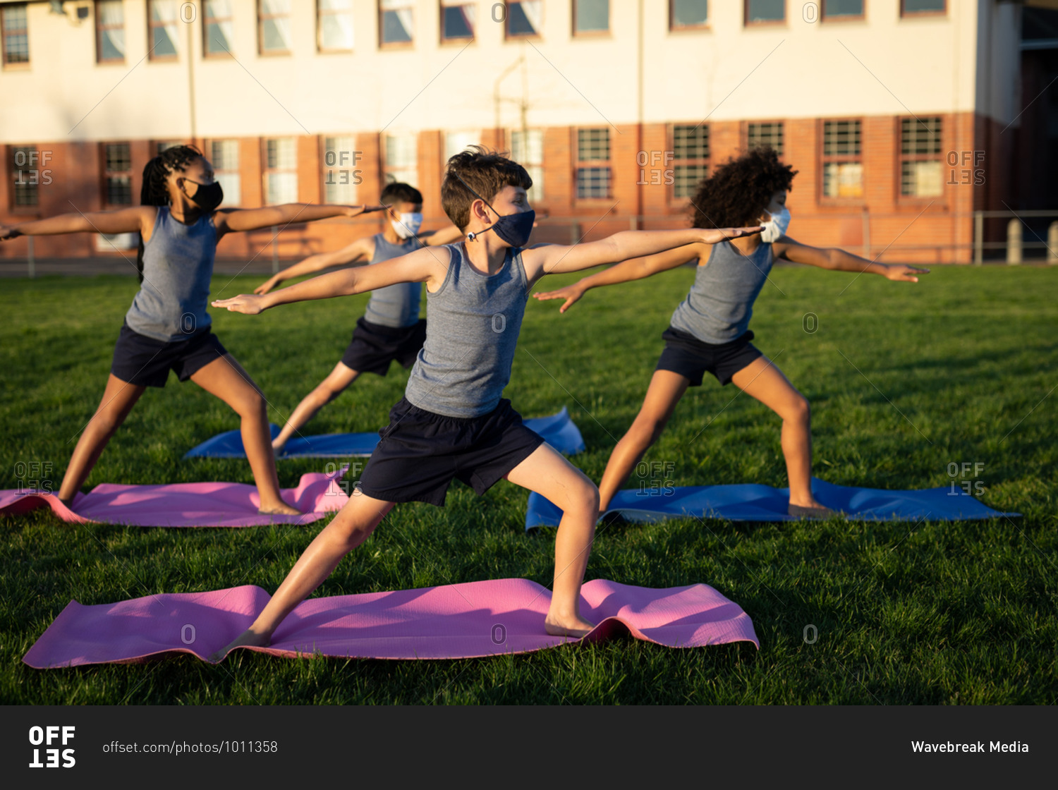 Group of multi ethnic kids wearing face masks performing yoga in the school garden. Primary education social distancing health safety during Covid19 Coronavirus pandemic.