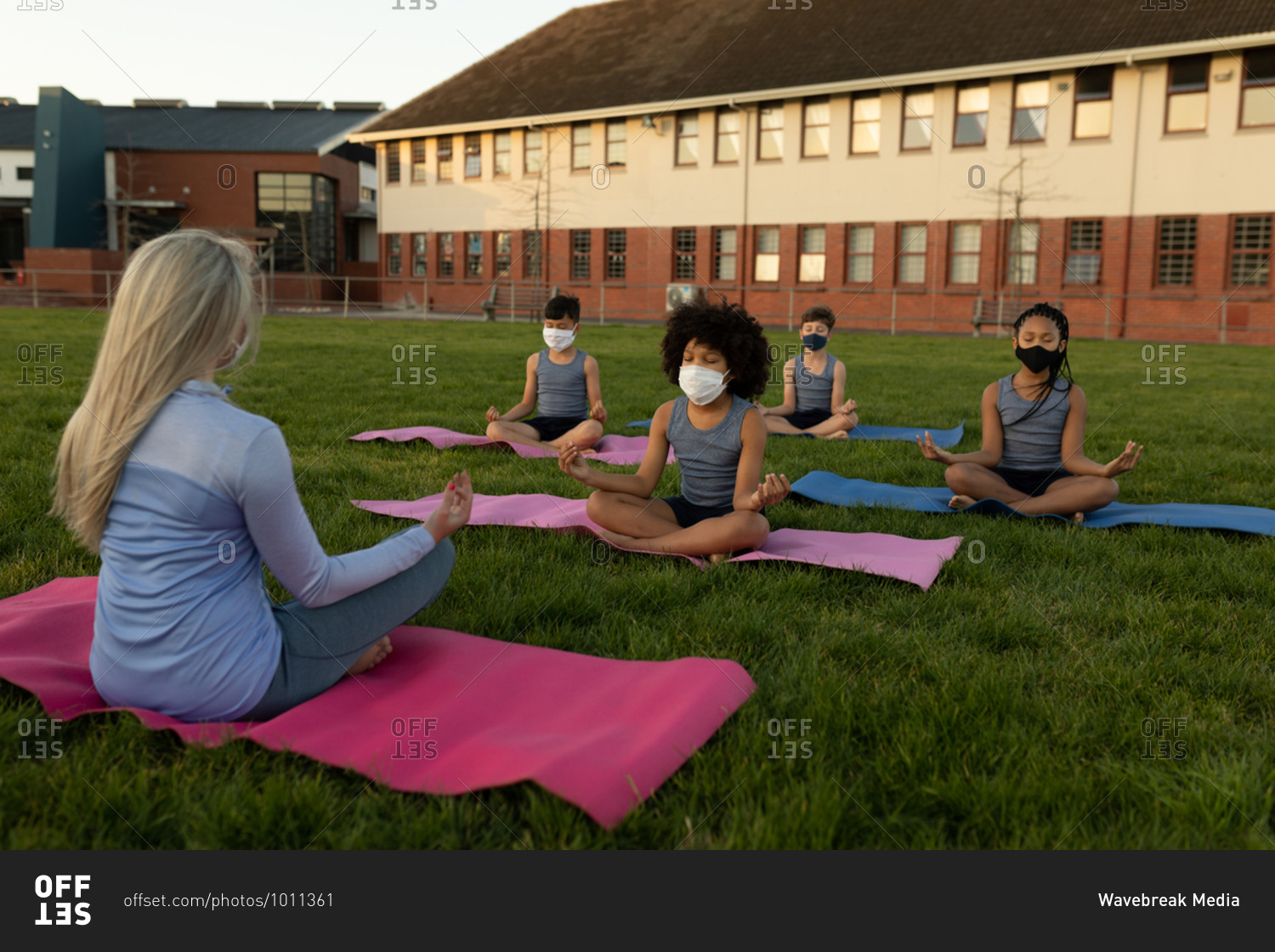 Group of multi ethnic kids and their female teacher wearing face masks performing yoga in the school garden. Primary education social distancing health safety during Covid19 Coronavirus pandemic.