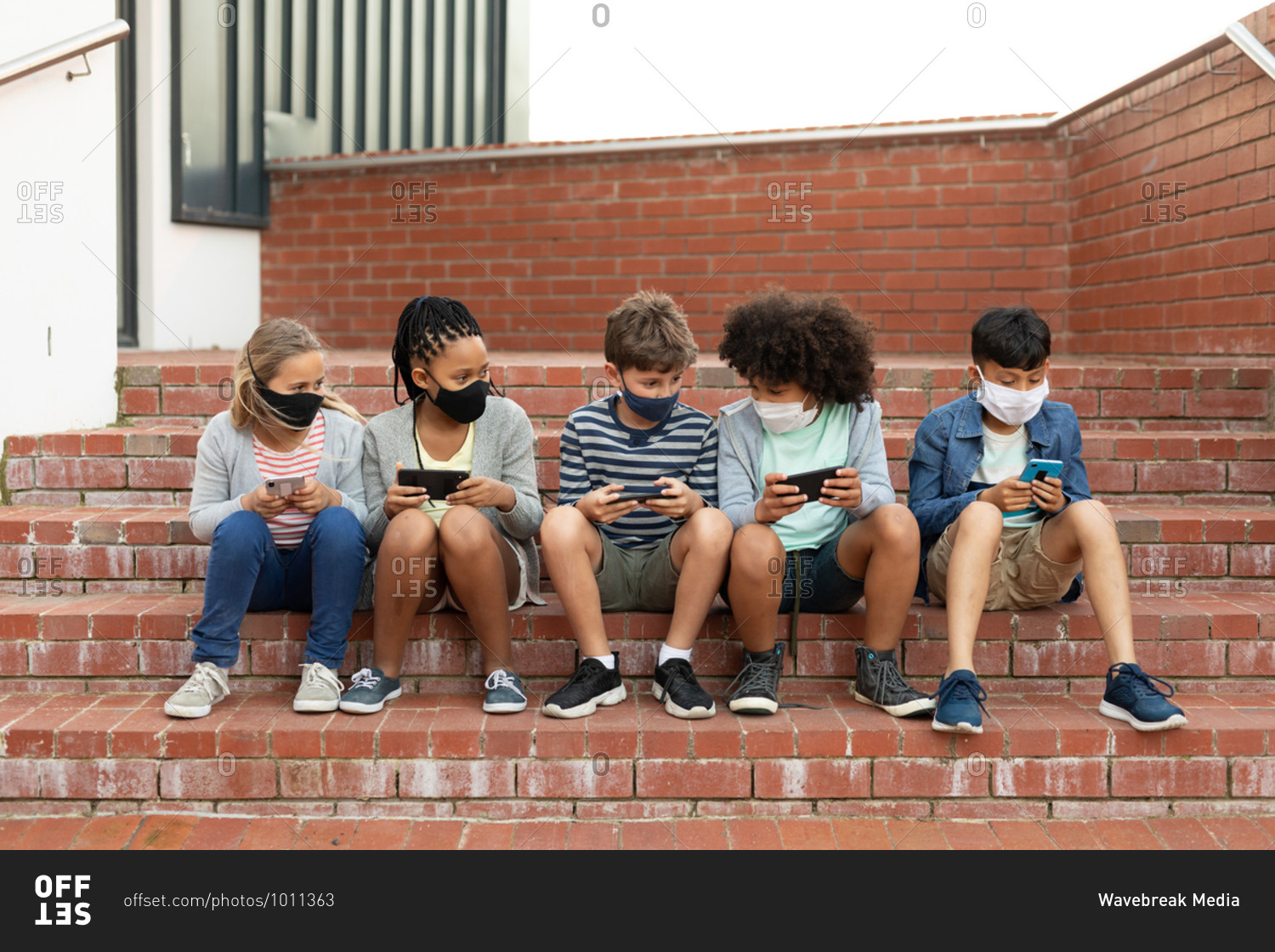 Group of multi ethnic kids wearing face masks using smartphones while sitting on the stairs during a break. Primary education social distancing health safety during Covid19 Coronavirus pandemic.