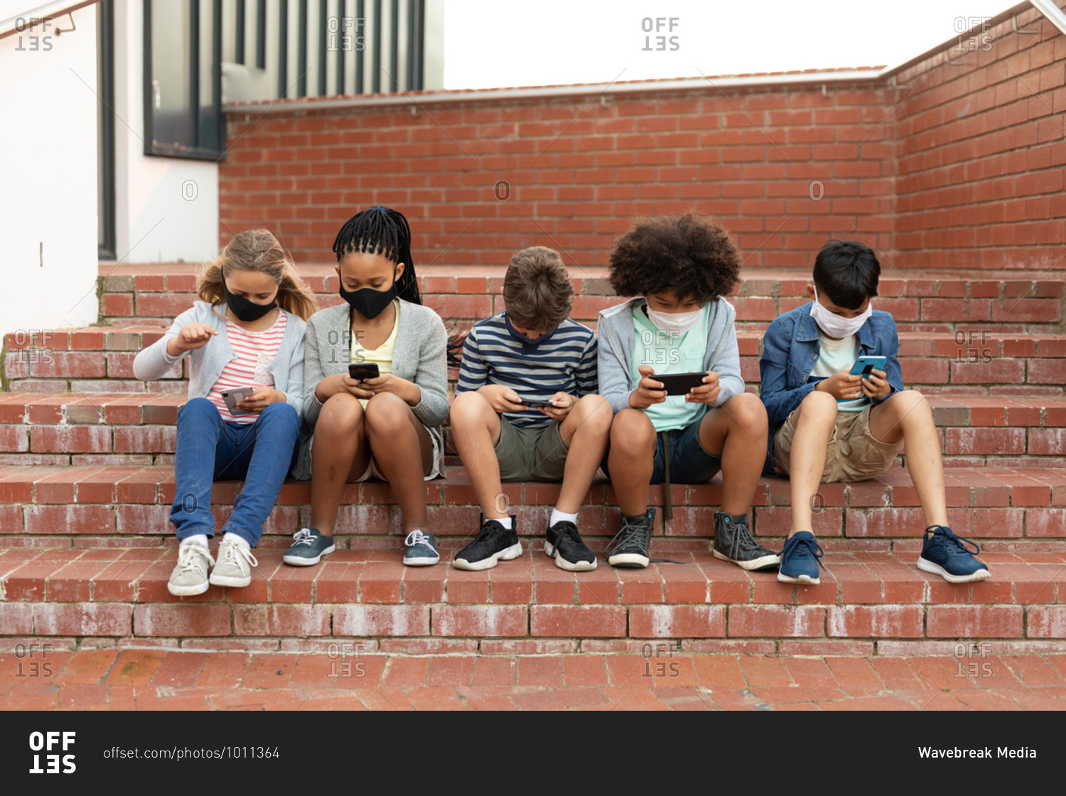 Group of multi ethnic kids wearing face masks using smartphones while sitting on the stairs during a break. Primary education social distancing health safety during Covid19 Coronavirus pandemic.