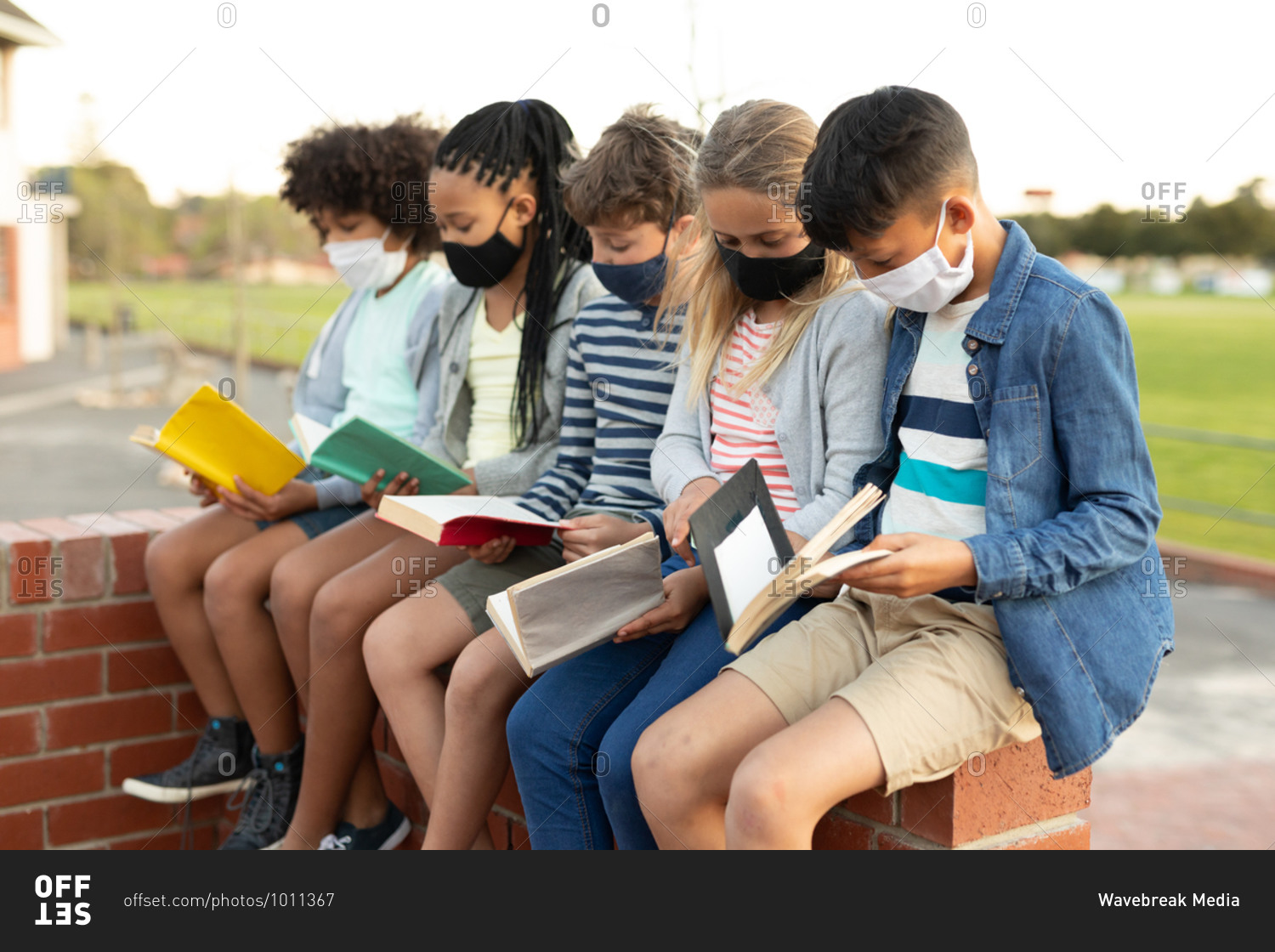 Group of multi ethnic kids wearing face masks reading books while sitting on the wall during a break. Primary education social distancing health safety during Covid19 Coronavirus pandemic.