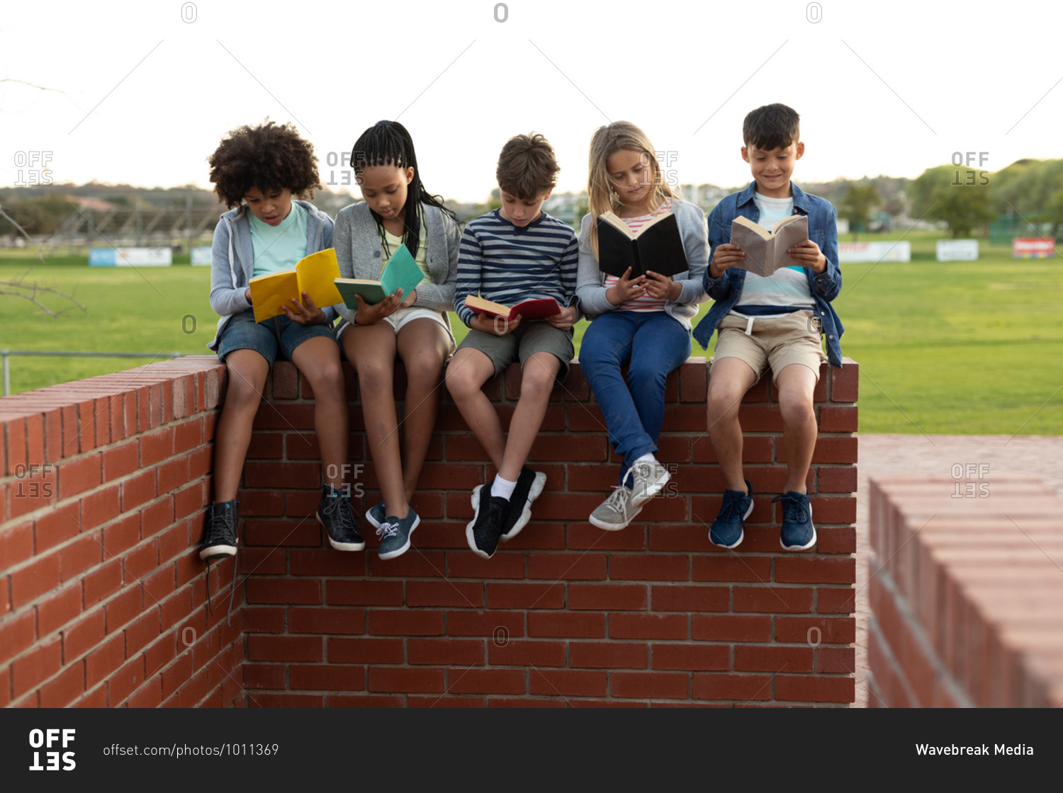 Group of multi ethnic kids reading books while sitting on the wall during a break. Primary education social distancing health safety during Covid19 Coronavirus pandemic.