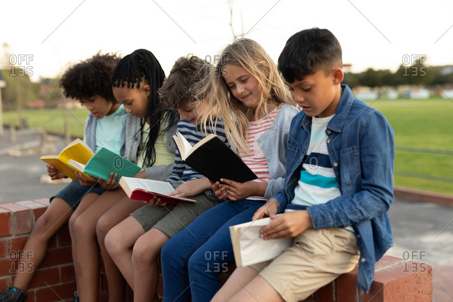 Group of multi ethnic kids reading books while sitting on the wall during a break. Primary education social distancing health safety during Covid19 Coronavirus pandemic.