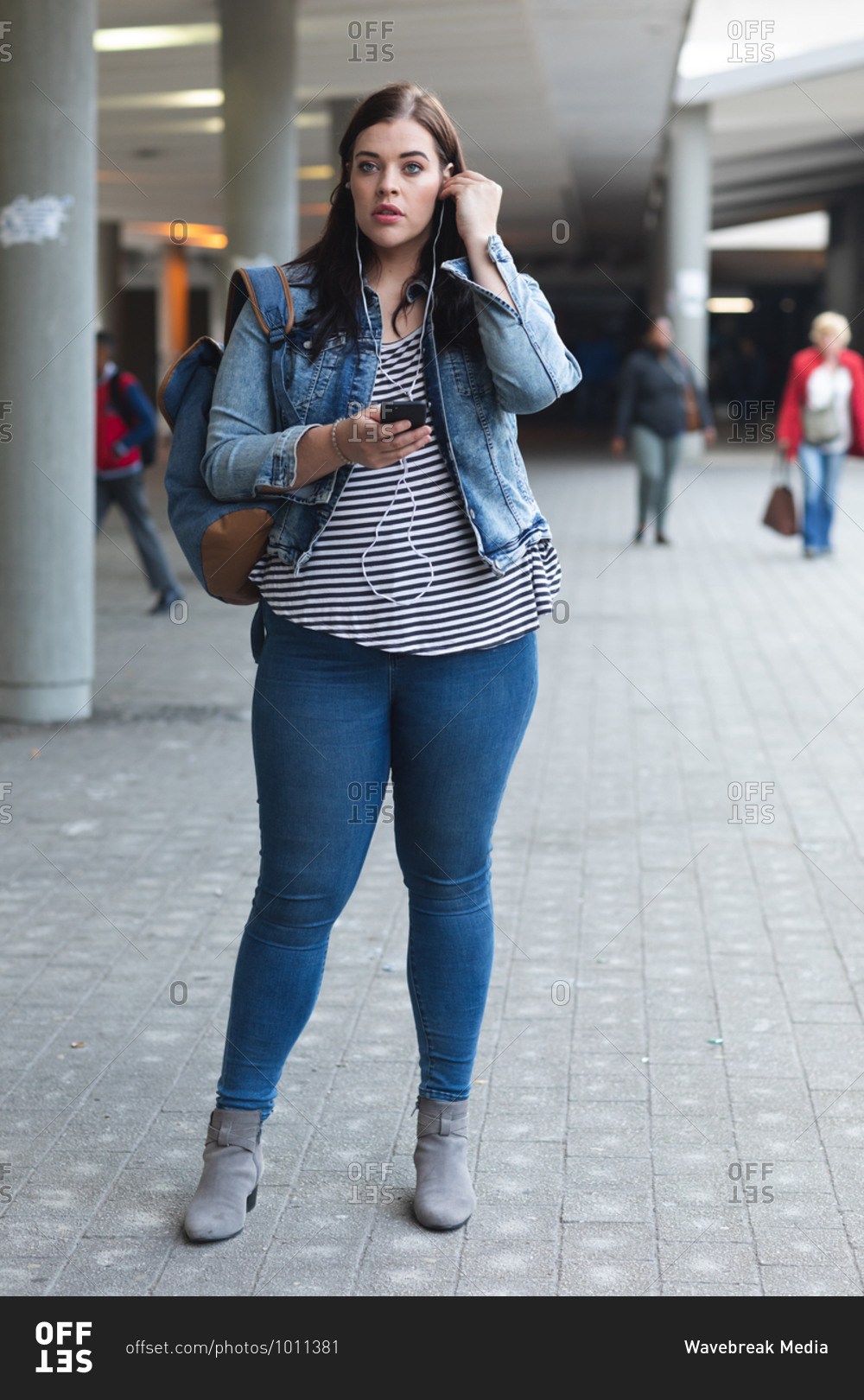 Curvy Caucasian woman out and about in the city streets during the day, smiling and using her smartphone wearing earphones, with modern building in the background