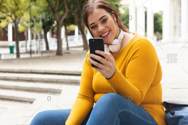Curvy Caucasian woman out and about in the city streets during the day, sitting on steps, smiling and using her smartphone wearing headphones with a historical building in the background