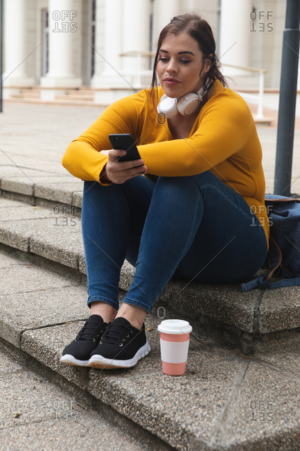 Curvy Caucasian woman out and about in the city streets during the day, sitting on steps with a takeaway coffee, using her smartphone, wearing headphones, with a historical building in the background