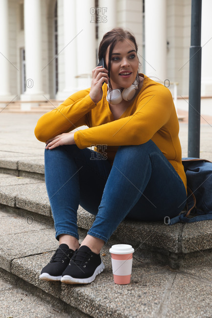 Curvy Caucasian woman out and about in the city streets during the day, sitting on steps with a takeaway coffee, talking on smartphone, wearing headphones, with a historical building in the background
