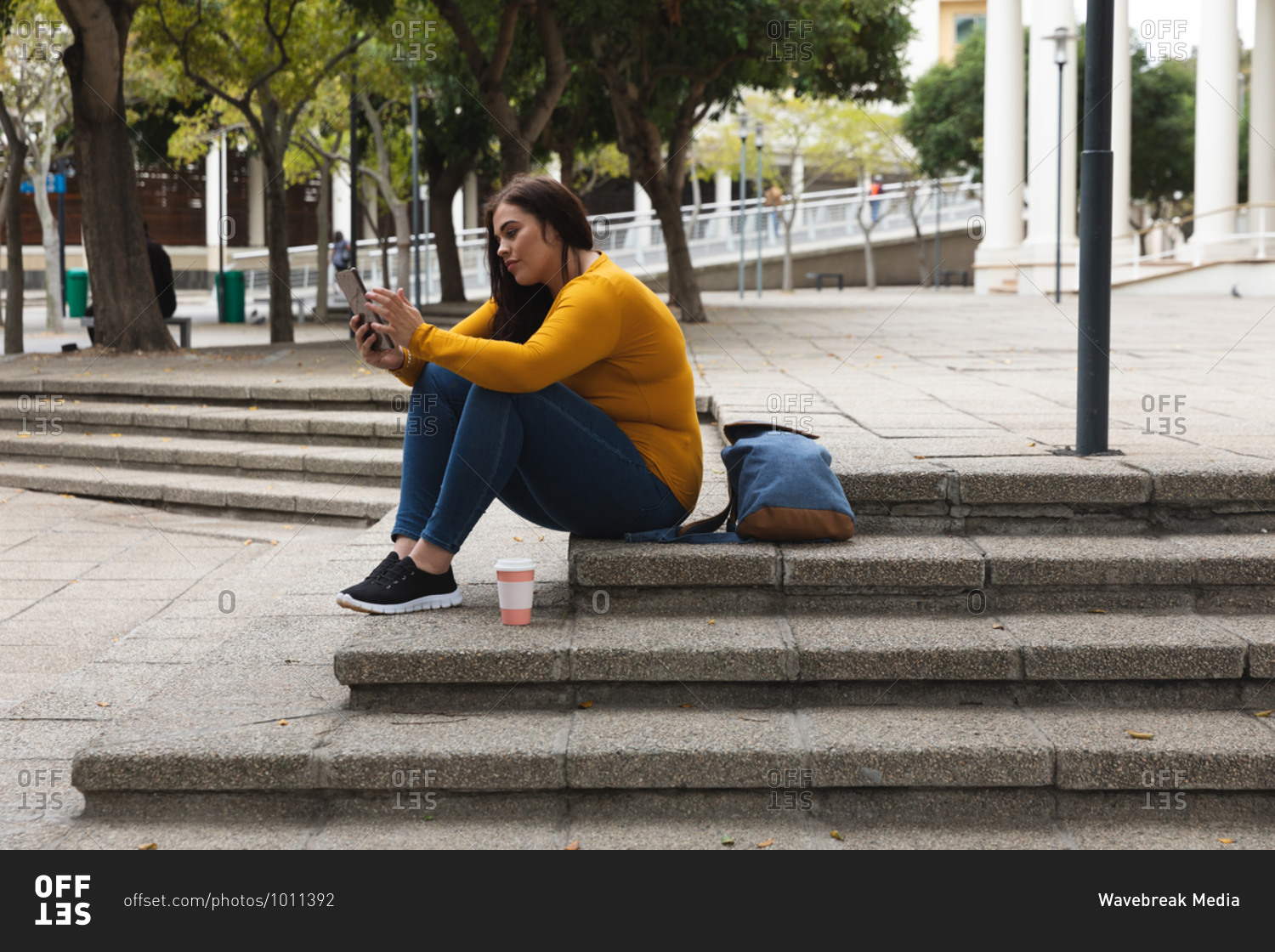 Curvy Caucasian woman out and about in the city streets during the day, sitting on steps with a takeaway coffee and her backpack, using a digital tablet, with a historical building in the background