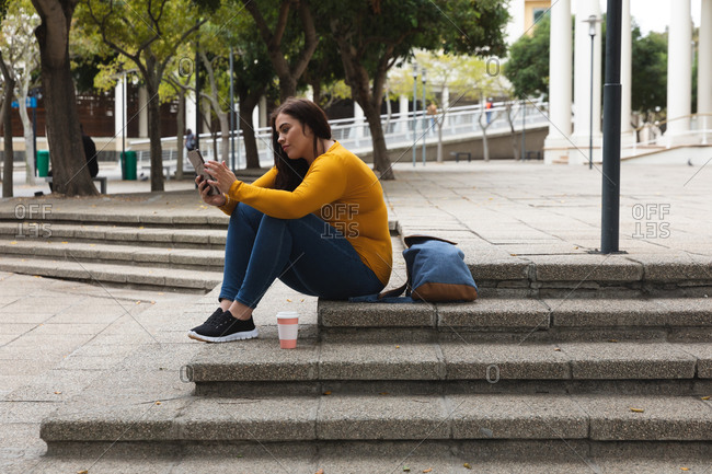 Curvy Caucasian woman out and about in the city streets during the day, sitting on steps with a takeaway coffee and her backpack, using a digital tablet, with a historical building in the background