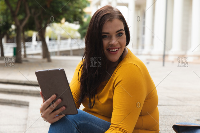 Portrait of a curvy Caucasian woman out and about in the city streets during the day, sitting on steps using a digital tablet and smiling to camera, with a historical building in the background
