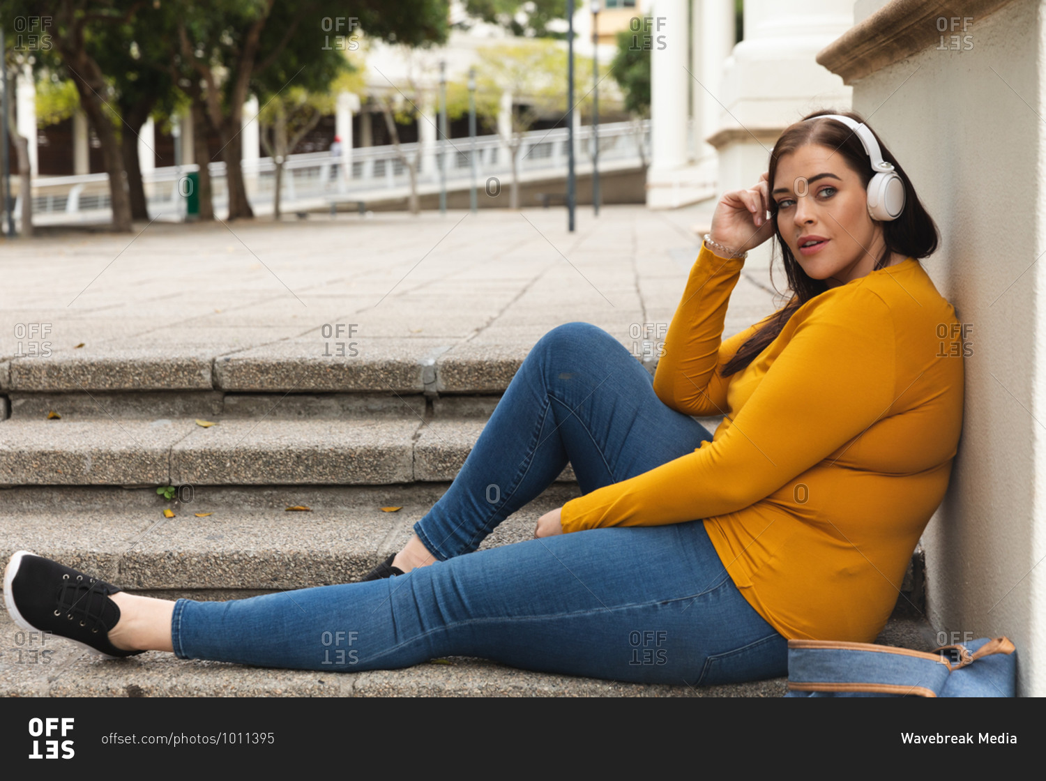 Curvy Caucasian woman out and about in the city streets during the day, sitting on steps, leaning back and listening to music on headphones, with historical building in the background
