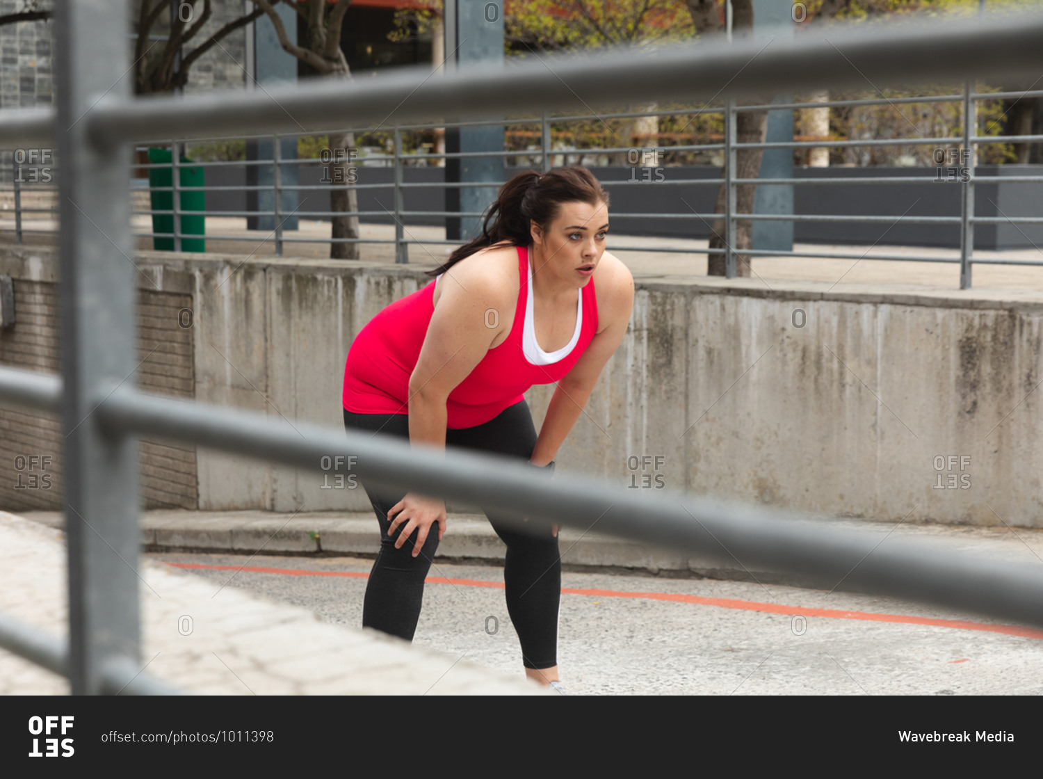Curvy Caucasian woman with long dark hair wearing sports clothes exercising in a city, leaning forward to take a rest during her work out, with modern buildings in the background