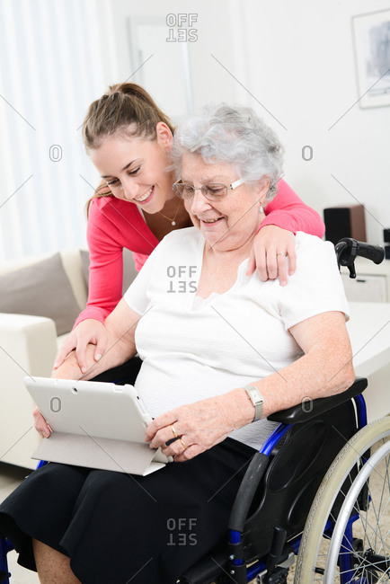 Cheerful young girl playing on internet with tablet computer and sharing time with old senior woman on wheelchair