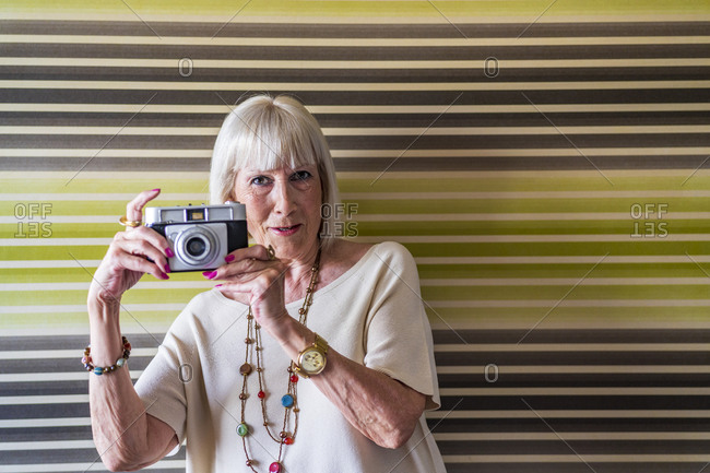 Stylish senior woman photographing with camera while standing against wall at home