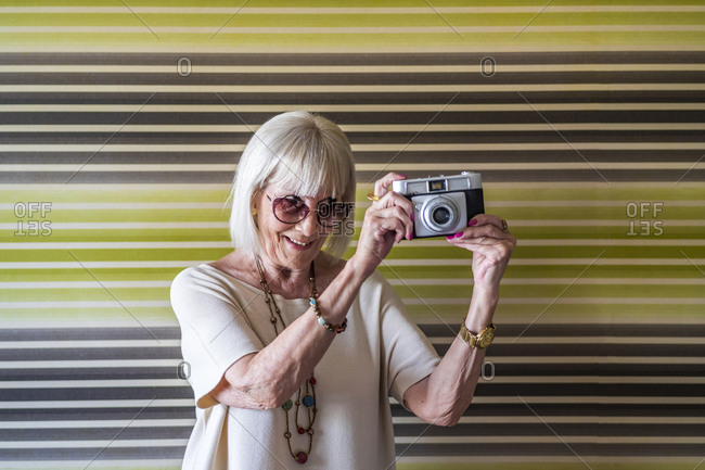 Stylish senior woman wearing sunglasses photographing with camera against wall at home