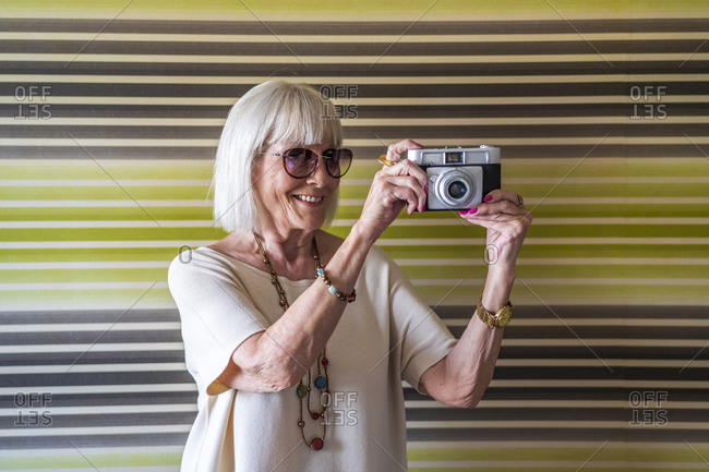 Smiling senior woman wearing sunglasses photographing with camera against wall at home