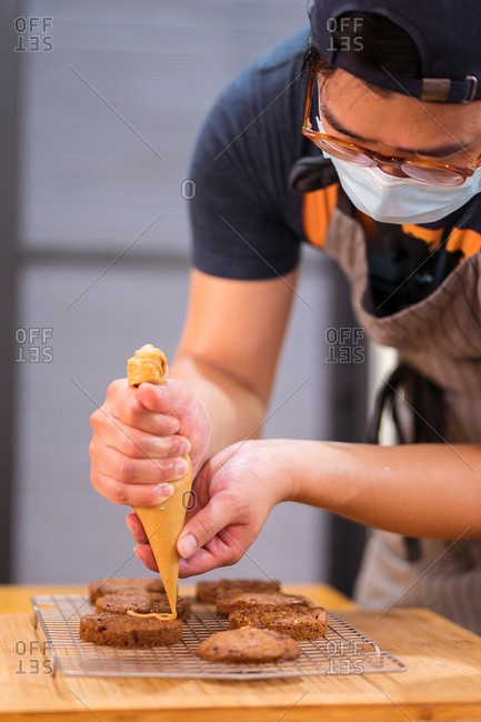 Focused Asian man in medical mask holding piping bag with caramel sauce topping chocolate chip cookies on cooling rack