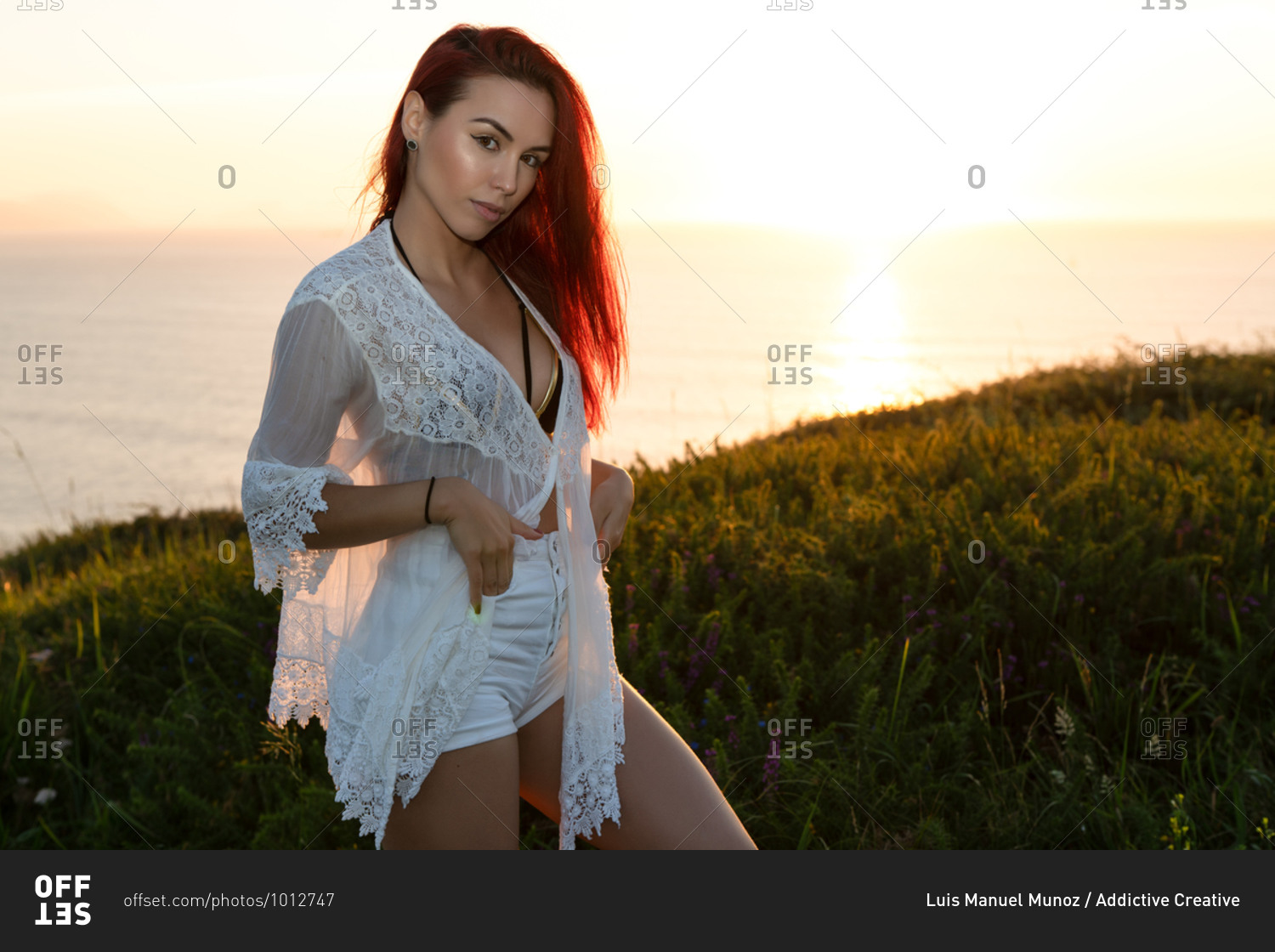 Alluring young red haired female in white translucent blouse and shorts looking at camera while standing on green meadow against calm sea during summer sunset