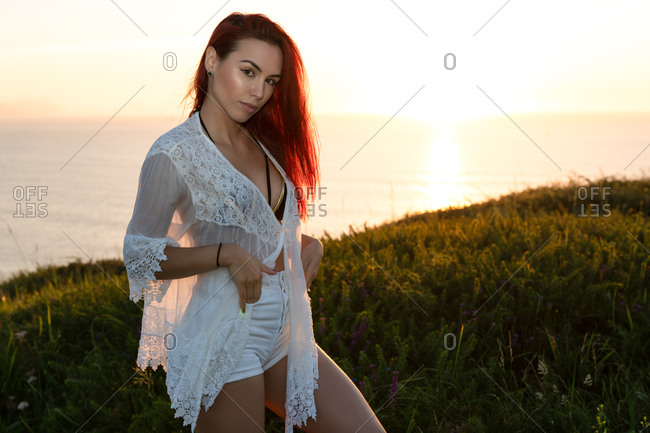 Alluring young red haired female in white translucent blouse and shorts looking at camera while standing on green meadow against calm sea during summer sunset