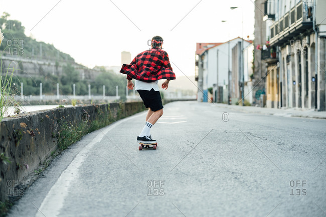 Back view of male hipster in checkered shirt riding longboard along asphalt road while entertaining during weekend in city