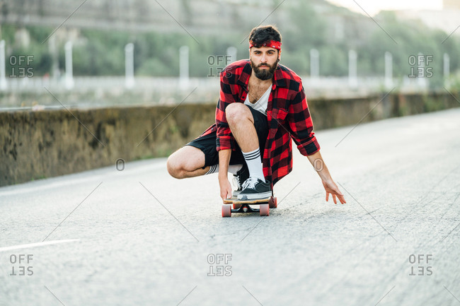 Male hipster in plaid shirt sitting on longboard and riding along road