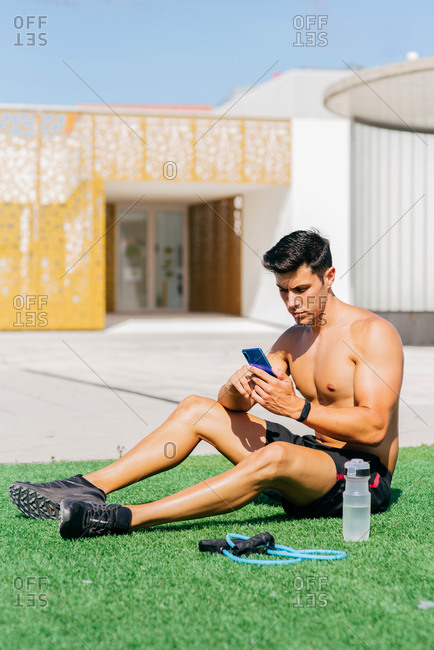 Determined athletic male with naked torso resting on lawn and browsing cellphone during training on sunny day
