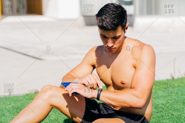 Athletic male with muscular torso sitting on grass and checking pulse on fitness tracker during workout on terrace in summer