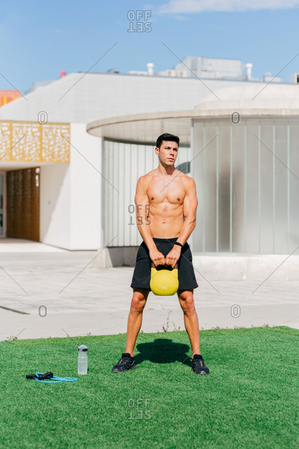 Handsome muscular shirtless sportsman standing on lawn and exercising with heavy kettlebell while looking away