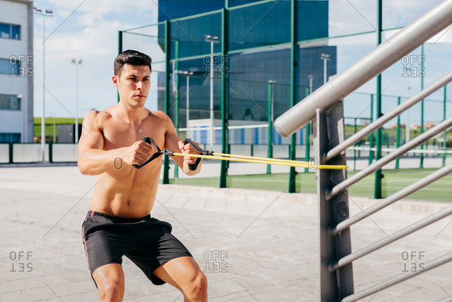 Focused shirtless male athlete doing exercises with elastic band during training in city in summer