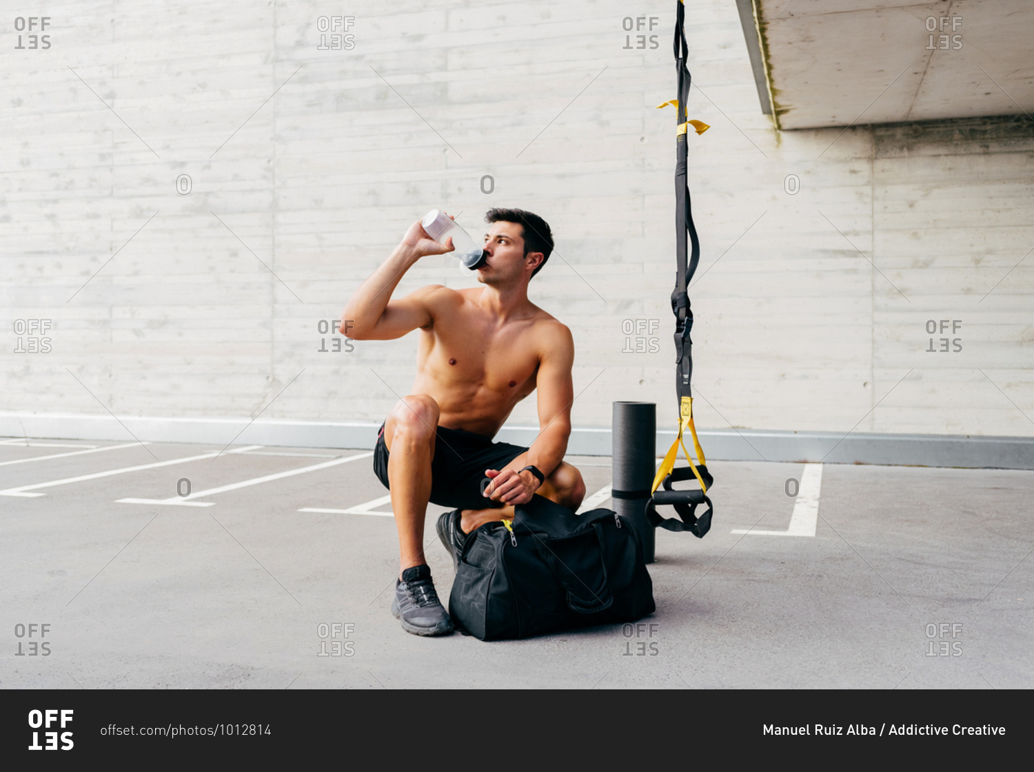 Relaxed sportsman with naked muscular torso crouching on street and drinking clean water from bottle after workout in city