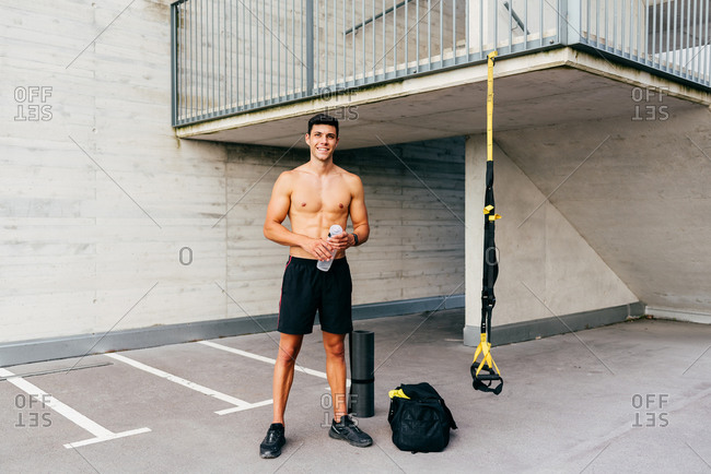 Relaxed happy sportsman with naked muscular torso standing on street holding water bottle resting after workout in city looking at camera