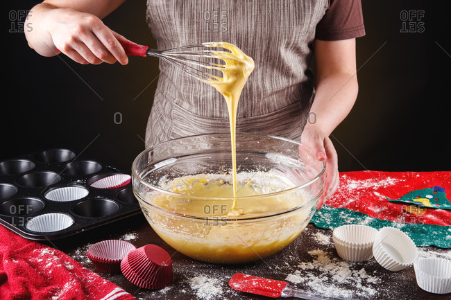 Crop anonymous housewife with whisk mixing dough in glass bowl while making delicious sweet cupcakes for Christmas celebration