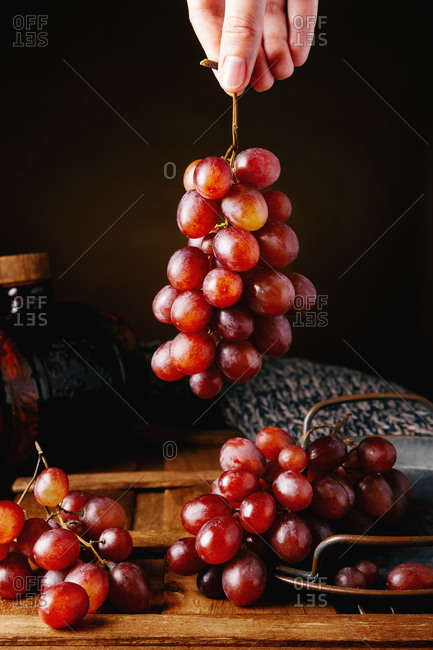 Crop unrecognizable person taking bunch of fresh juicy red grapes from tray placed on wooden table against black background