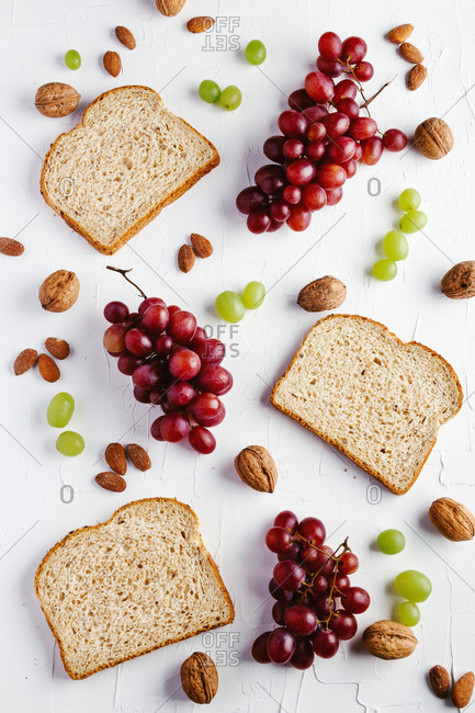 Top view composition with sliced bread and red and green grapes arranged on white table with walnuts and almonds
