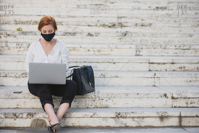 Businesswoman in formal wear and protective mask sitting on stone steps in city and working on netbook during COVID 19 epidemic