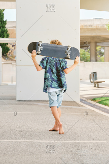 Vertical photo of the back of a boy with a printed shirt and shorts carrying a skateboard on his back while walking down the street