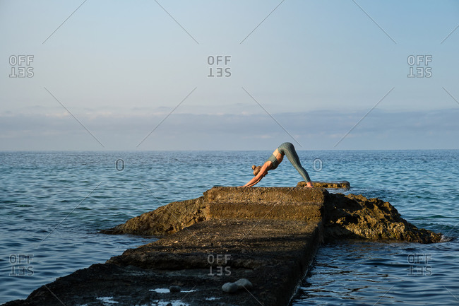 Side view of distant unrecognizable woman standing in Downward Facing Dog position while practicing yoga on rocky pier at seaside