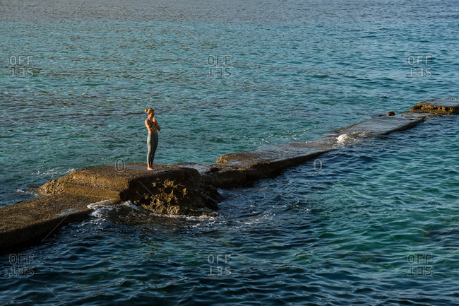 Full body side view of distant female in sportswear standing in Mountain pose with prayer hands and meditating during yoga practice on rocky pier at seaside