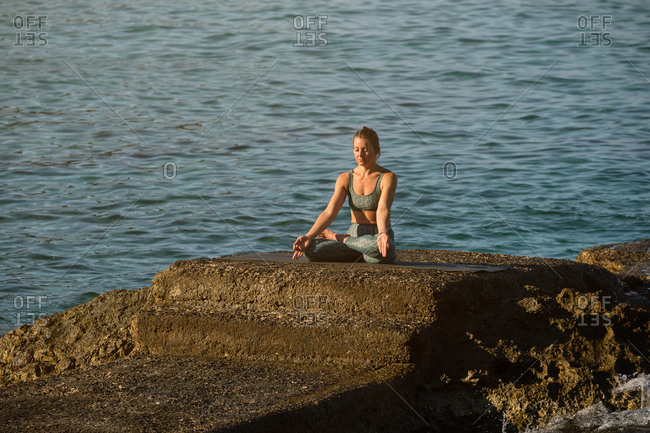 Peaceful female sitting on stone in Padmasana and meditating with mudra gesture near sea during sundown