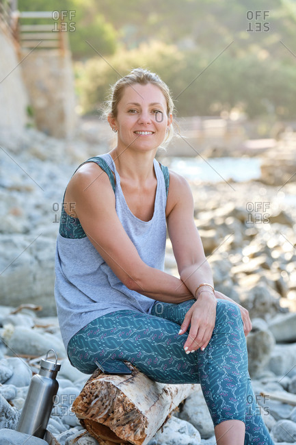 Smiling female in sportswear sitting on tree trunk while resting after meditation on rocky seashore and looking at camera