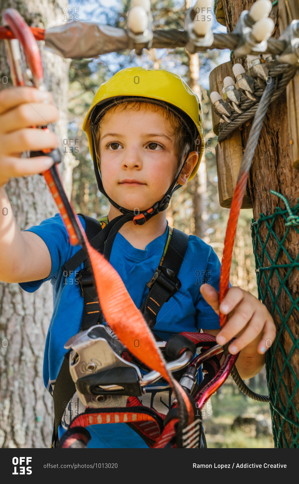 Adorable child in protective helmet and safety harness putting carabine on rope while spending weekend in adventure park in summer