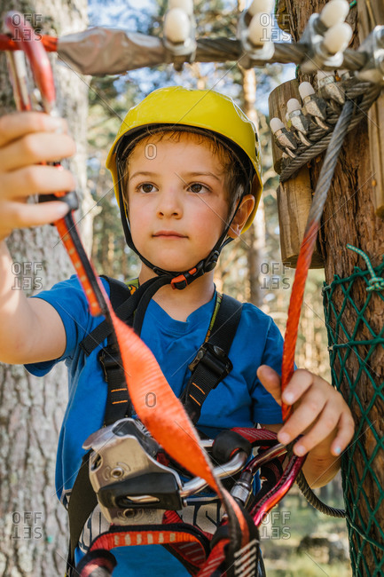 Adorable child in protective helmet and safety harness putting carabine on rope while spending weekend in adventure park in summer